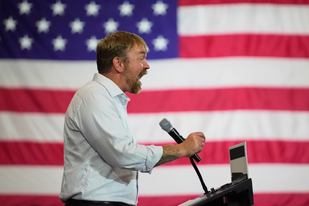 Graham Platner stands at a podium. He appears to be shouting into a microphone. A large American flag takes up the background.